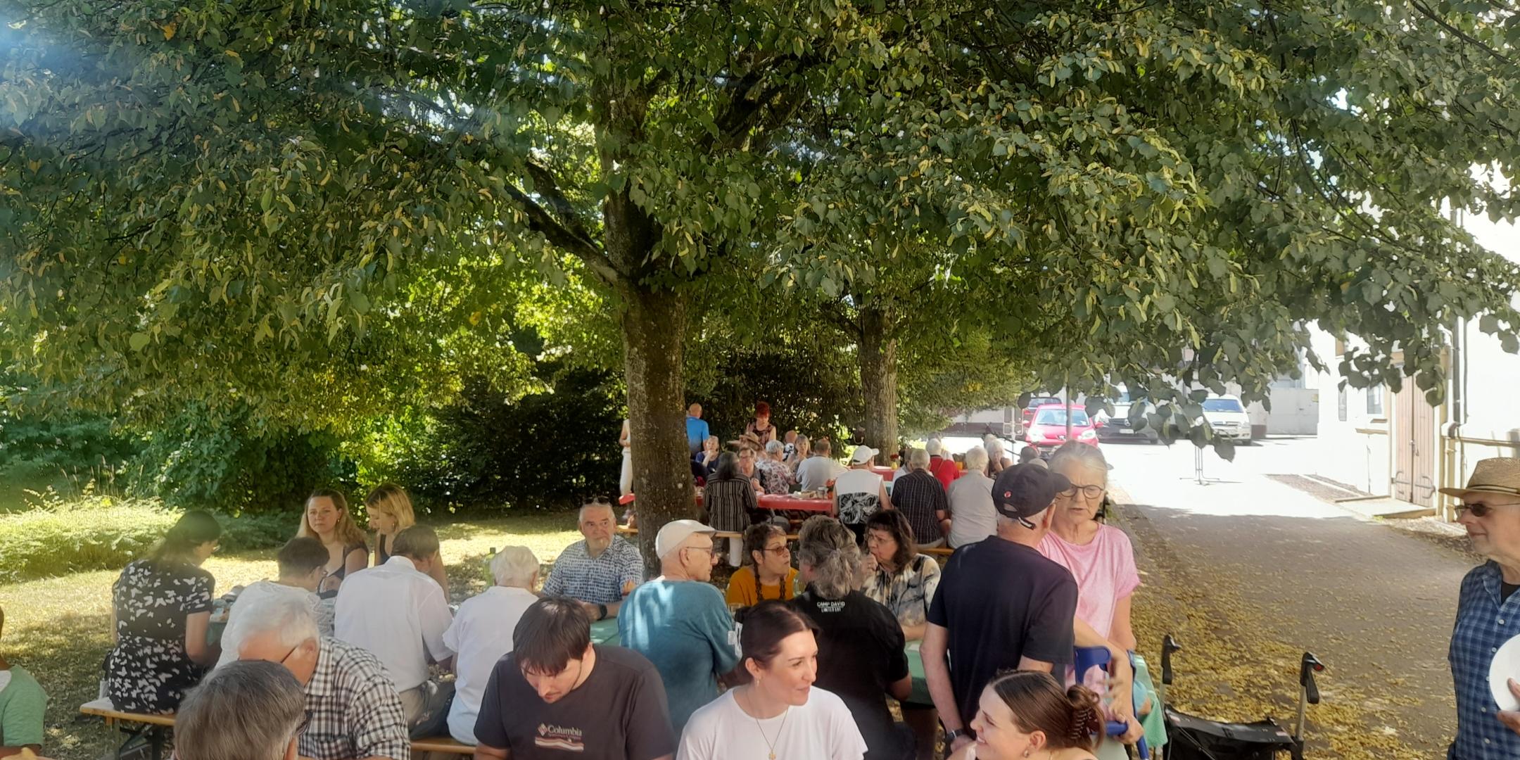 Unter den Bäumen sitzen Menschen  an Tischen, essen und unterhalten sich