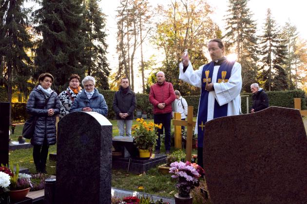 Gemeinsam am Grab an Allerheiligen Ein Priester im liturgischen Gewand steht auf dem Friedhof vor einem Grab und segnet es mit erhobenen Arm. Um ihn stehen einige Menschen.