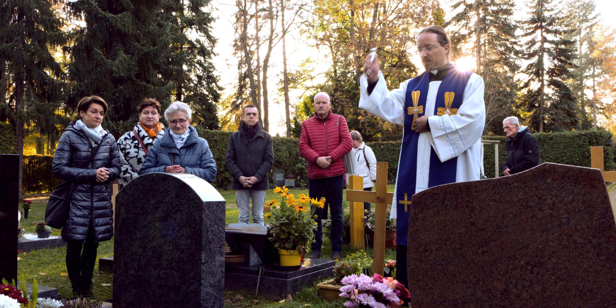Zusammen am Grab zur Gräbersegnung Ein Priester im liturgischen Gewand steht auf dem Friedhof vor einem Grab und segnet es mit erhobenen Arm. Um ihn stehen einige Menschen.