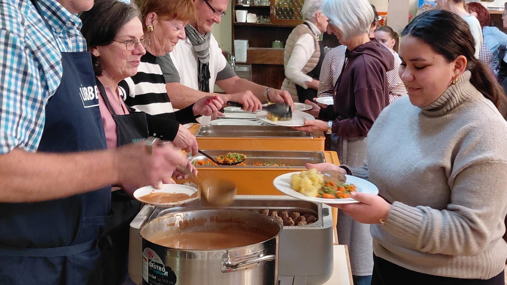 Ein Mann mit Schürze schöpft aus einem Kessel. Ihm gegenüber eine weibliche Jugendliche mit einem Teller