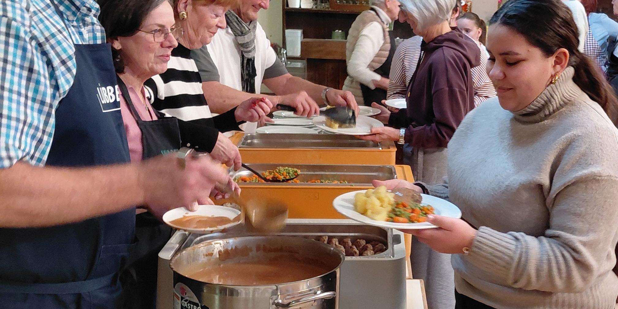 Ein Mann mit Schürze schöpft aus einem Kessel. Ihm gegenüber eine weibliche Jugendliche mit einem Teller