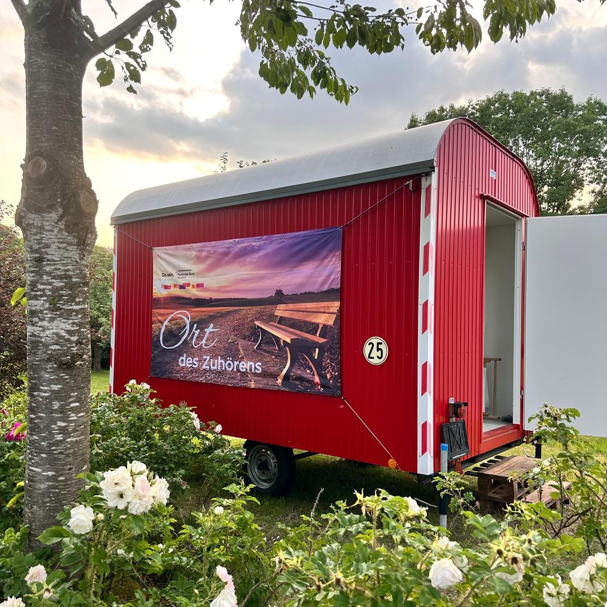 Ein Roter Bauwagen mit der Aufschrift 'Ort des Zuhörens' inmitten von blühenden Büschen und einem Baum