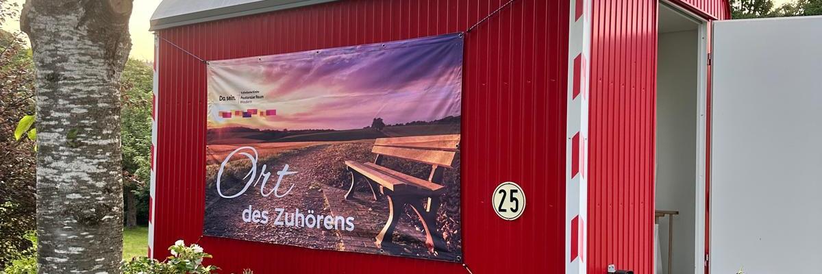 Ein Roter Bauwagen mit der Aufschrift 'Ort des Zuhörens' inmitten von blühenden Büschen und einem Baum
