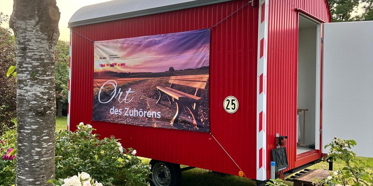 Ein Roter Bauwagen mit der Aufschrift 'Ort des Zuhörens' inmitten von blühenden Büschen und einem Baum