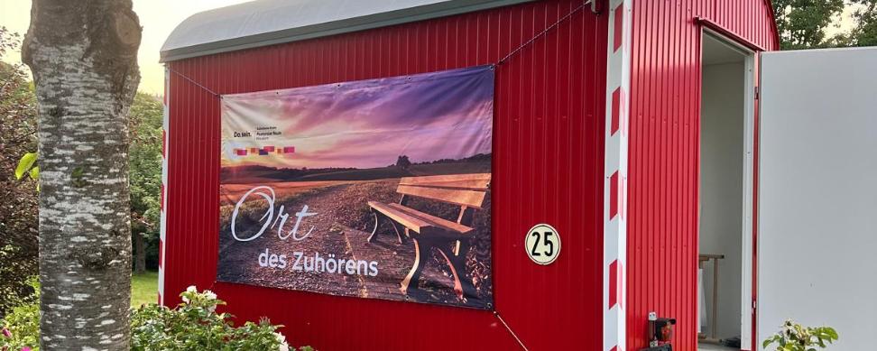 Ein Roter Bauwagen mit der Aufschrift 'Ort des Zuhörens' inmitten von blühenden Büschen und einem Baum