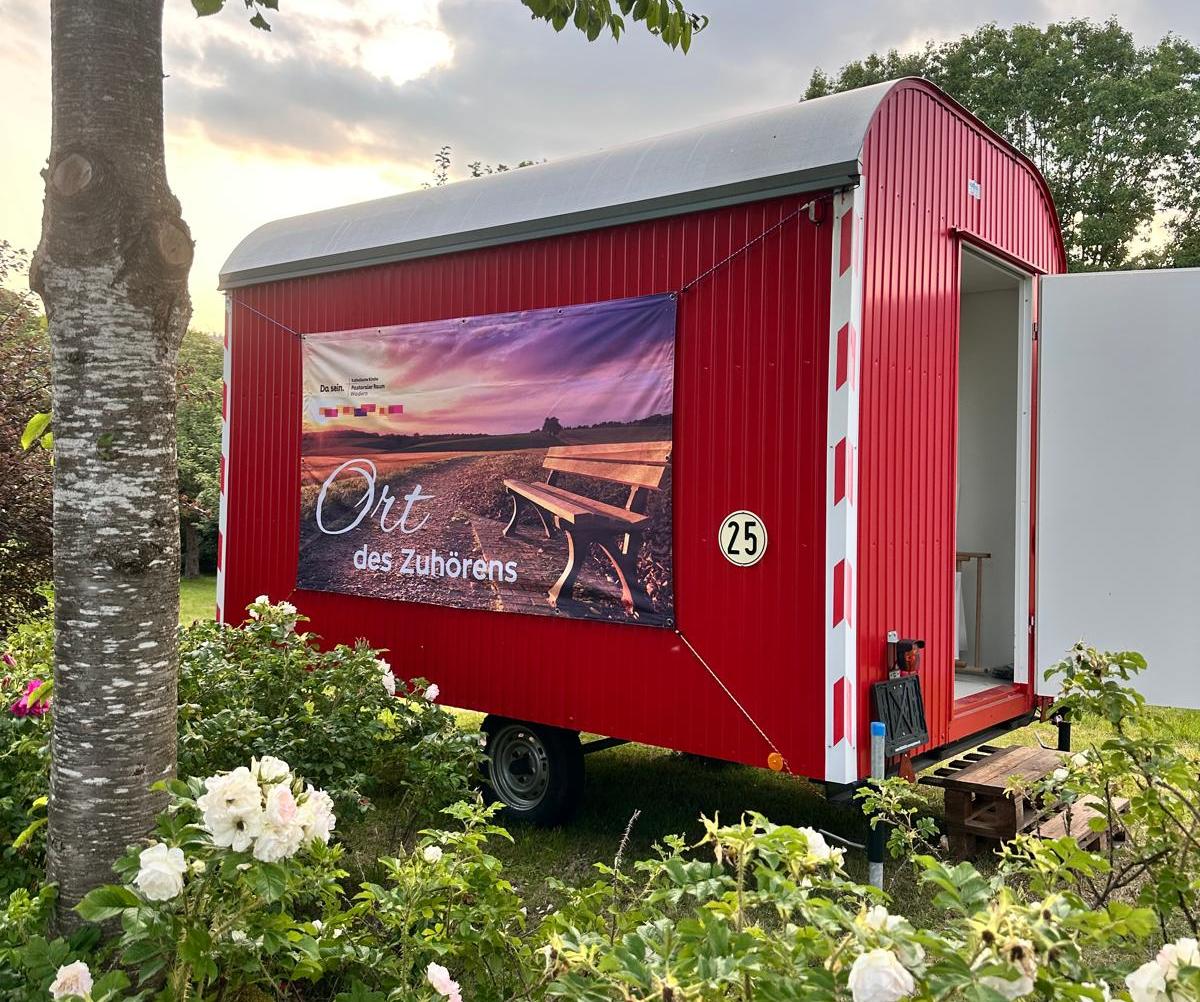 Ein Roter Bauwagen mit der Aufschrift 'Ort des Zuhörens' inmitten von blühenden Büschen und einem Baum