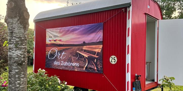 Ein Roter Bauwagen mit der Aufschrift 'Ort des Zuhörens' inmitten von blühenden Büschen und einem Baum
