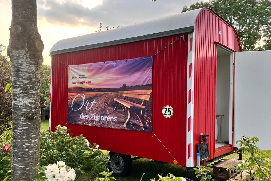 Ein Roter Bauwagen mit der Aufschrift 'Ort des Zuhörens' inmitten von blühenden Büschen und einem Baum