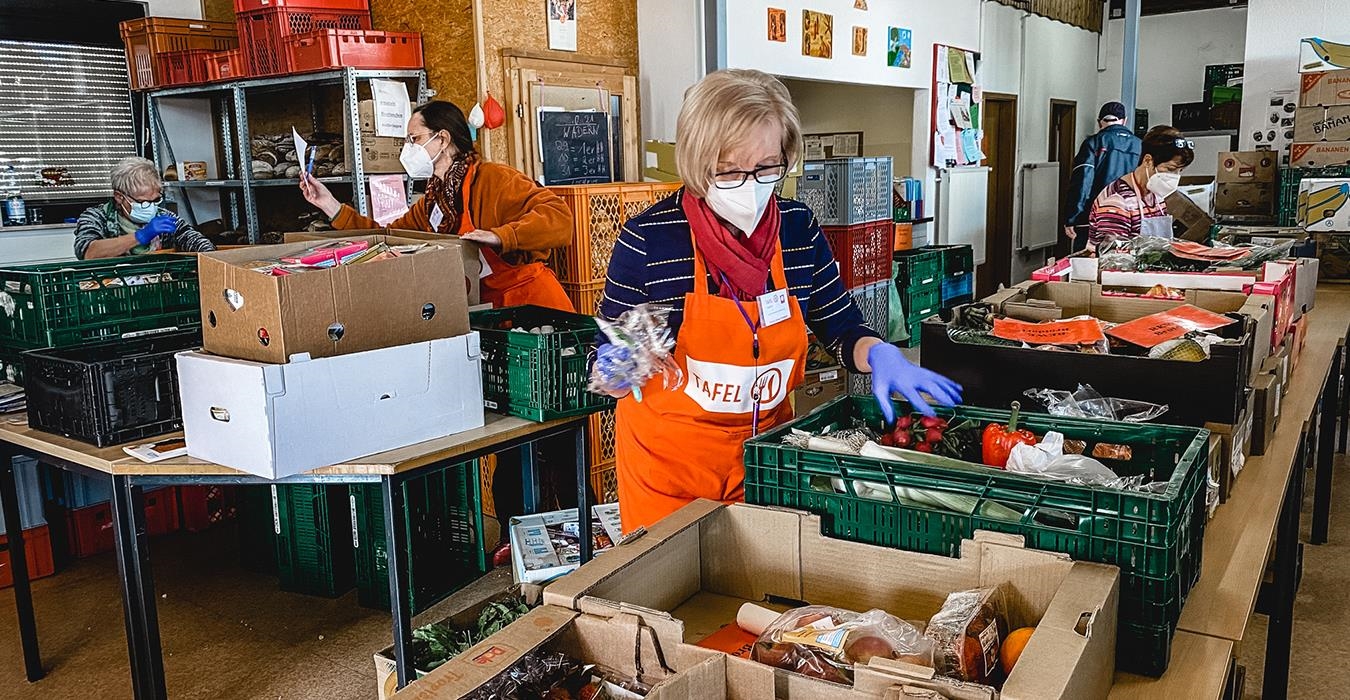 Inmitten Regalen von Lebensmitteln stehen zwei Menschen, die sortieren. Sie tragen Schützen mit der Aufschrift Tafel