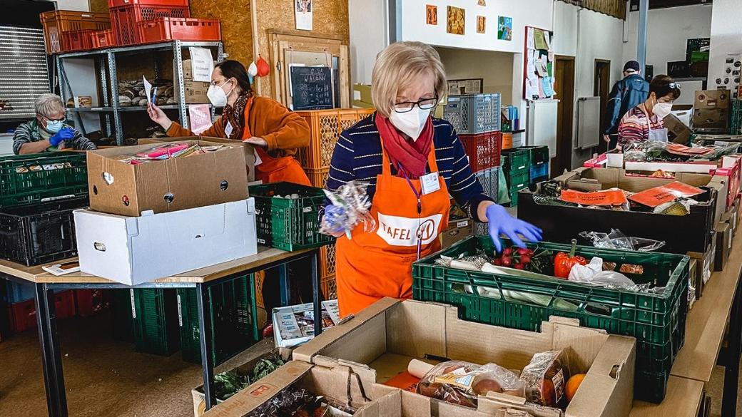 Inmitten Regalen von Lebensmitteln stehen zwei Menschen, die sortieren. Sie tragen Schützen mit der Aufschrift Tafel