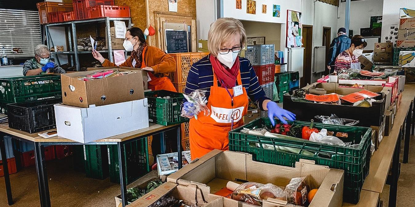 Inmitten Regalen von Lebensmitteln stehen zwei Menschen, die sortieren. Sie tragen Schützen mit der Aufschrift Tafel