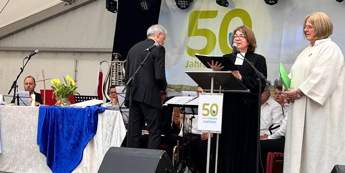 Man sieht eine Bühne. Im Hintergrund des Musikverein Losheim. Im Vordergrund zwei Frauen, die den Gottesdienst leiten.