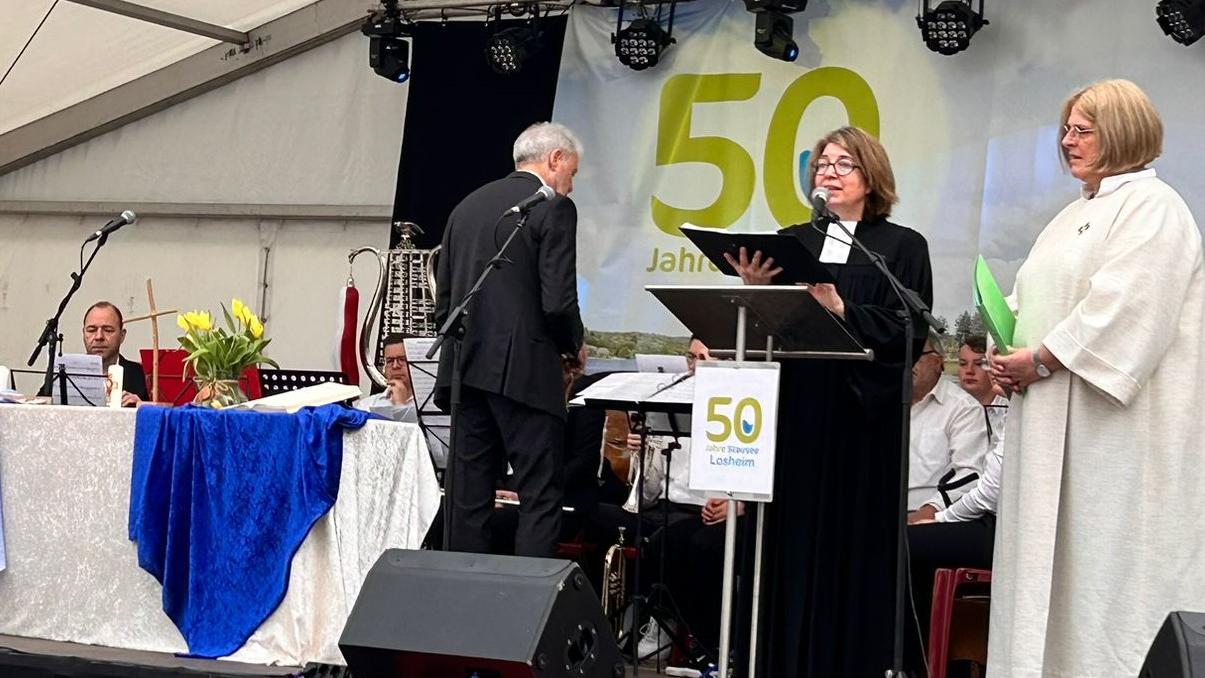 Man sieht eine Bühne. Im Hintergrund des Musikverein Losheim. Im Vordergrund zwei Frauen, die den Gottesdienst leiten.