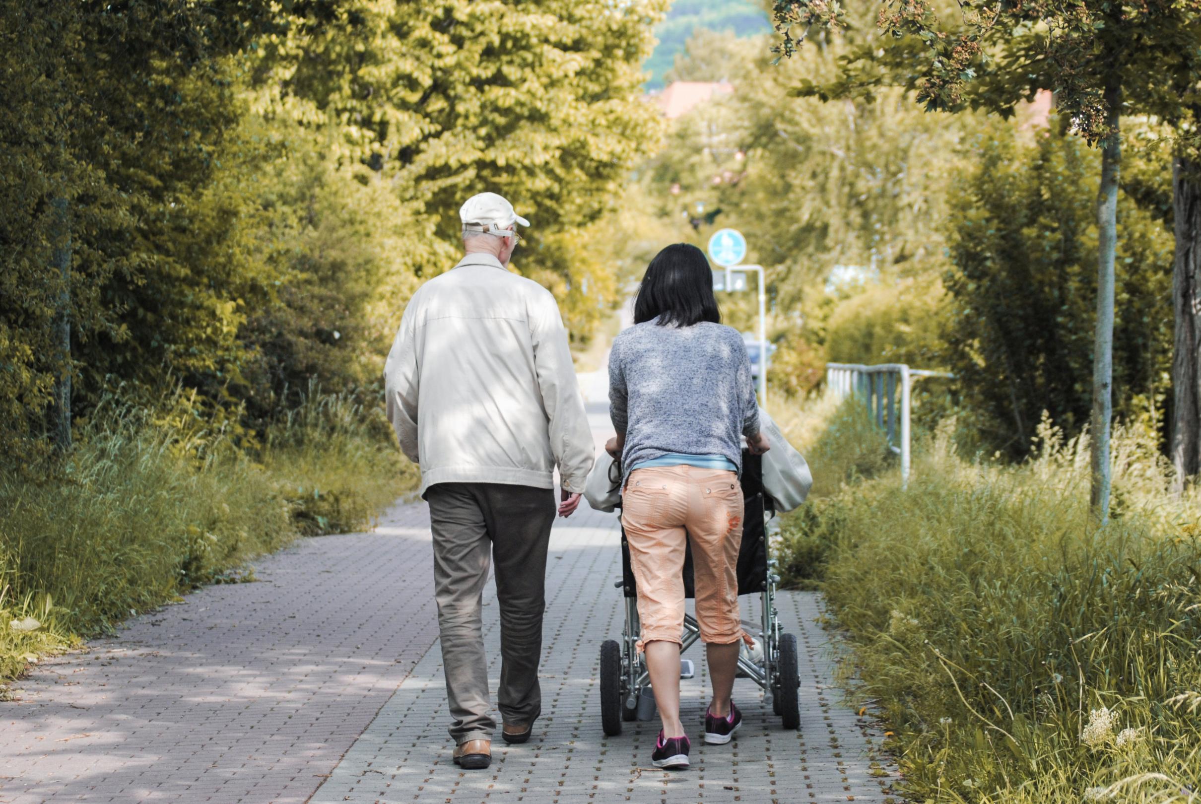Eine Frau schiebt einen Rollstuhl durch einen Park. Neben ihr geht ein älterer Herr.