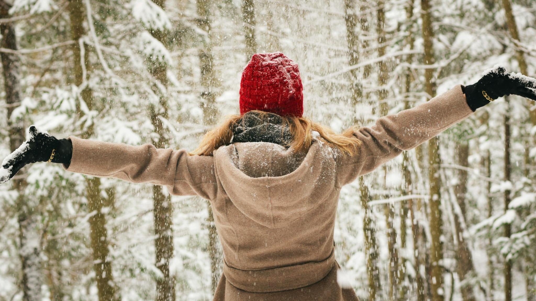 Eine junge Frau steht in Winterkleidern mit ausgebreiteten Armen vor der Winterlandschaft